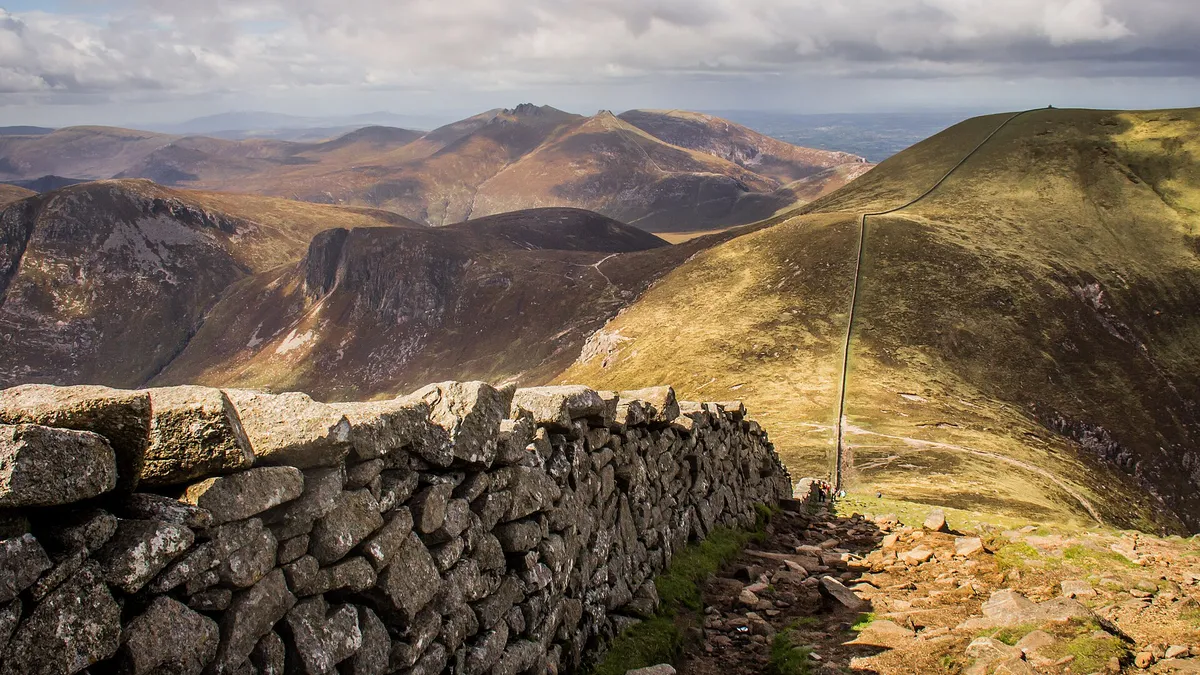 The Mourne Wall running along a grassy ridge, with the peaks of the Mourne Mountains behind it