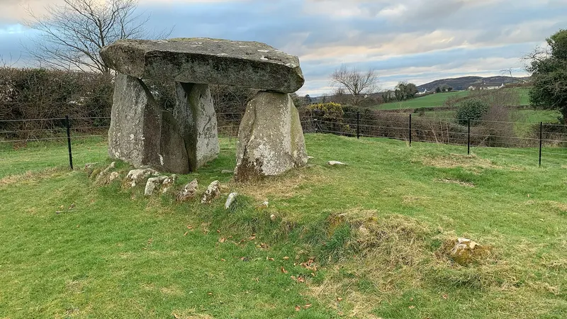 Ballykeel Dolmen, a capstone on three upright stones, at the end of a low cairn in a green field