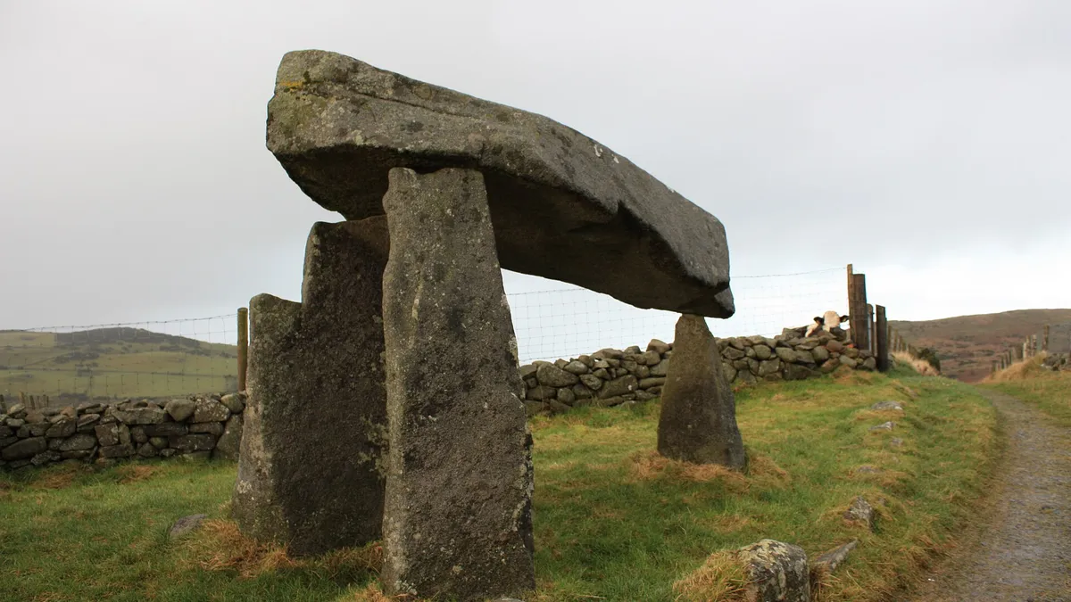 The Legananny Dolmen, a flat capstone raised on three tall stones, beside a stone wall and track