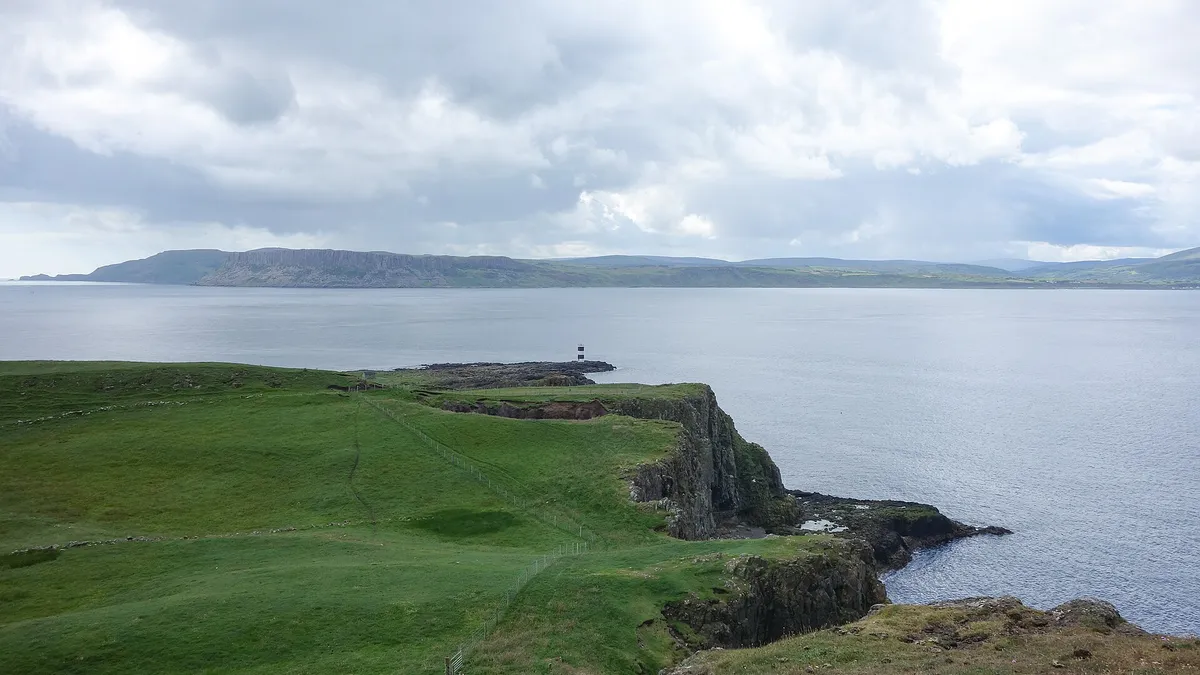 Green clifftops on Rathlin Island running out to a small lighthouse, with the mainland across the water