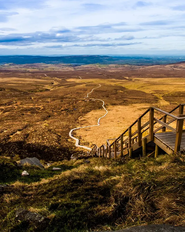 The wooden stairway of the Cuilcagh boardwalk climbing a slope, with brown bog and a lake spread out below