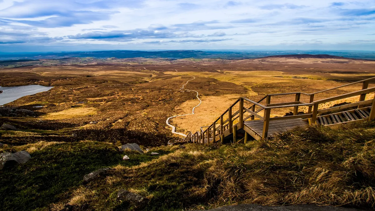 The wooden stairway of the Cuilcagh boardwalk climbing a slope, with brown bog and a lake spread out below