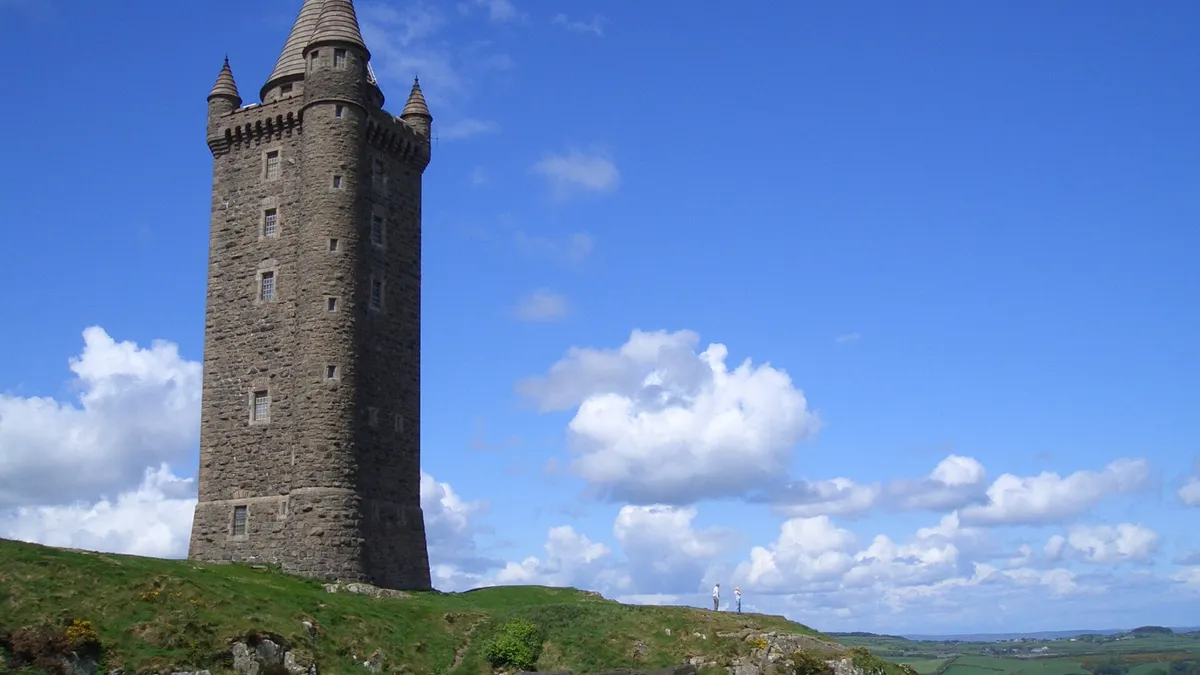 Scrabo Tower, a tall stone tower with a pointed roof, on a green hilltop under a blue sky