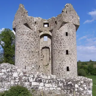 The two round towers of Monea Castle rising above a low stone wall under a blue sky