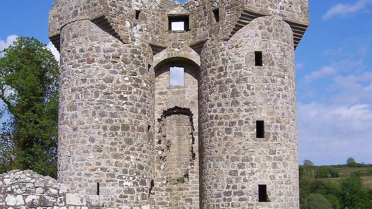 The two round towers of Monea Castle rising above a low stone wall under a blue sky
