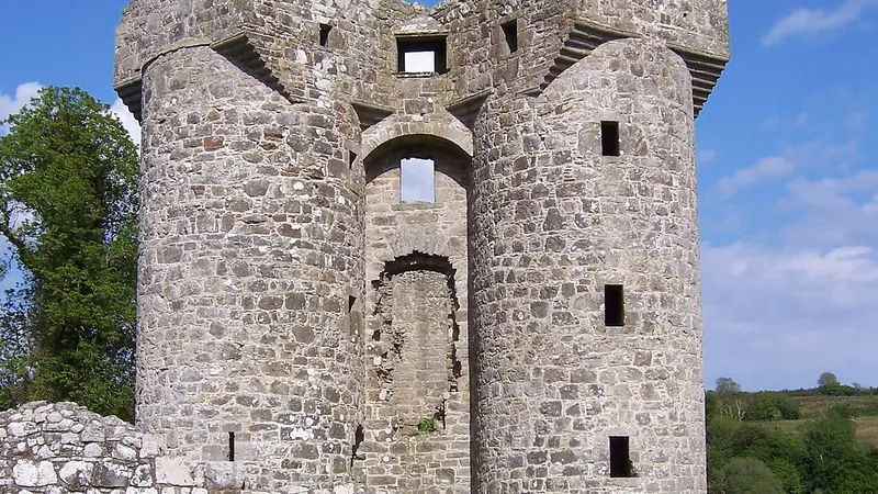 The two round towers of Monea Castle rising above a low stone wall under a blue sky