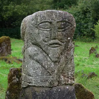 The two faced carved stone figure in Caldragh graveyard, lichen on its face, with old grave markers behind it