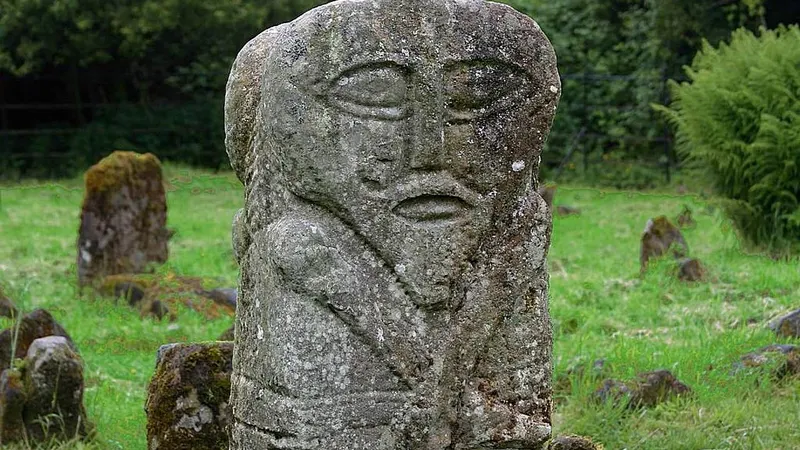 The two faced carved stone figure in Caldragh graveyard, lichen on its face, with old grave markers behind it