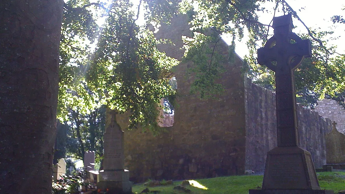 The gable of a ruined stone church at Killevy among trees, with a stone cross in the graveyard