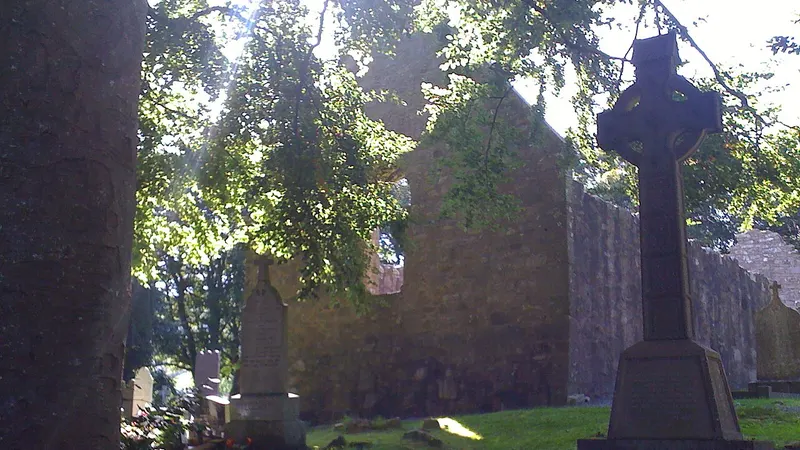 The gable of a ruined stone church at Killevy among trees, with a stone cross in the graveyard