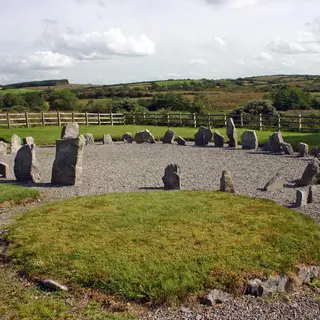 A ring of low standing stones on gravel beside a grassy cairn, with farmland behind