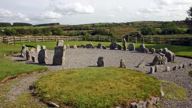 A ring of low standing stones on gravel beside a grassy cairn, with farmland behind