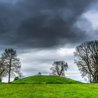 The grassy mound of Navan Fort on a hilltop, with bare trees against a stormy sky