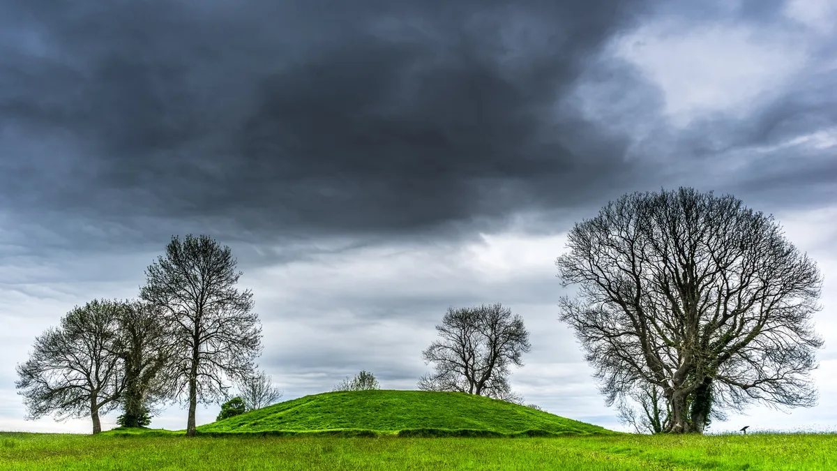 The grassy mound of Navan Fort on a hilltop, with bare trees against a stormy sky