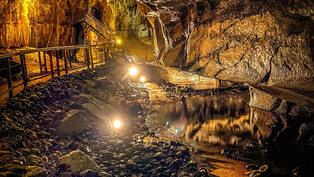 A lit walkway running beside dark water between cave walls and hanging formations in the Marble Arch Caves