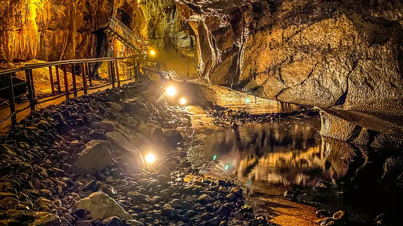 A lit walkway running beside dark water between cave walls and hanging formations in the Marble Arch Caves